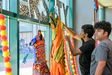 Dwóch mężczyzn wiesza kolorowe stroje na wieszaku przy oknie. Jeden trzyma pomarańczowy strój z zielonymi akcentami. W tle dekoracje z kwiatów i mozaikowy napis. Za oknem widoczne rowery i budynki.