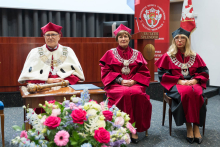 Trzy osoby w togach akademickich siedzą na krzesłach. Osoba po lewej w białej todze z berłem, pozostałe w czerwonych togach i biretach. Przed nimi stół z bukietem kwiatów. W tle baner z herbem uczelni i napisem „Veritatis Splendor”.