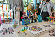 Stół z białym obrusem w ludowe wzory, na którym ustawiono rękodzieła: figurkę kota z rybą, kolorowego ptaka, niebieską komodę z kwiatami i inne ozdoby. W tle osoby przy oknie i dekoracje na szybie.