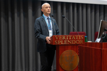 Mężczyzna, prof. Niemczyk, w ciemnym garniturze i niebieskim krawacie stoi za drewnianą mównicą z napisem „VERITATIS SPLENDOR” i emblematem z orłem. Na mównicy są dwa mikrofony. W tle szare zasłony, obok monitor.