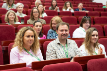 Grupa osób siedzi w czerwonych fotelach audytorium. Większość ma na szyjach zielone smycze z identyfikatorami. W tle widoczne są rzędy pustych i zajętych miejsc.