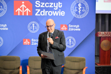 Mężczyzna w średnim wieku, dziekan wydziału, w ciemnym garniturze, białej koszuli i bordowym krawacie trzyma mikrofon oraz niebieską teczkę z logo Instytutu Ochrony Zdrowia. W tle niebieska ścianka z logotypami Instytutu i napisem „Szczyt Zdrowie”.