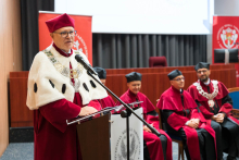 Rektor w czerwonym stroju ceremonialnym z białym kołnierzem i łańcuchem rektorskim przemawia do mikrofonu. W tle cztery osoby w czerwonych togach i biretach, dwa czerwone banery z logotypem uczelni i wydziału oraz drewniana mównica.