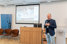 A man in a blue jacket stands at the lectern and holds a microphone. Behind him a screen with a presentation and a photo of the couple and the text: “Nutrition labeling on the front of the package - simplification or misinformation?”. Next to him is a blue banner with the words “Warsaw Medical University Faculty of Health Sciences.” In the foreground, three brown chairs.
