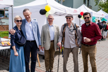 Pięć osób stoi przed białymi namiotami wystawowymi. Są to władze Wydziału Lekarskiego. Dwie osoby po lewej są elegancko ubrane, trzecia trzyma laskę i ma jasny garnitur. Czwarta ma kapelusz i plecak, piąta nosi czerwoną koszulę w kratę i trzyma kubek. W tle balony dekoracyjne.