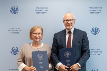 Kobieta, dyrektor NIO - po lewej i mężczyzna, rektor WUM po prawej - stoją na tle ścianki z logo Warszawskiego Uniwersytetu Medycznego. Obie trzymają granatowe teczki z herbem uczelni. Jedna osoba ubrana jest w jasny sweter, druga w ciemny garnitur, białą koszulę i bordowy krawat. Tło z powtarzającym się logo i nazwą uczelni.