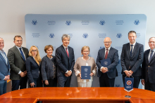 Dziewięć osób stoi w rzędzie przed niebieską ścianką z logotypami Warszawskiego Uniwersytetu Medycznego. Osoby są elegancko ubrane, dwie trzymają teczki z logo uczelni. Przed nimi znajduje się drewniany stół konferencyjny.