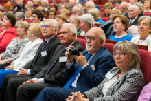 Grupa osób siedzi w audytorium podczas konferencji. Na pierwszym planie mężczyzna w niebieskim garniturze trzyma aparat, obok kobieta w marynarce w kratkę. W tle widoczni inni uczestnicy na czerwonych krzesłach. Słuchają z uwagą.