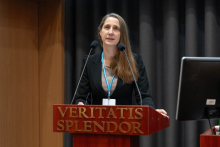 Kobieta, prof. Domitrz, z ciemnymi długimi włosami i w ciemnym stroju formalnym stoi za mównicą z napisem „VERITATIS SPLENDOR”. Na szyi ma identyfikator na niebieskiej smyczy. Przed nią znajdują się dwa mikrofony, a po prawej stronie widoczny jest monitor komputerowy. Tło stanowią ciemne zasłony.