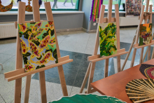 Trzy sztalugi z obrazami w pomieszczeniu. Lewy obraz przedstawia kolorowe motyle na zielonym tle, środkowy – liście tropikalnych roślin, a prawy częściowo widoczny motyw roślinny. Na pierwszym planie wachlarze i tkaniny, w tle okna.