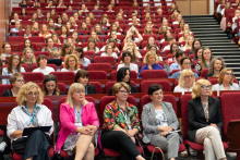Aula wypełniona uczestnikami siedzącymi w czerwonych fotelach. W pierwszym rzędzie pięć kobiet w różnych stylizacjach. W tle kolejne rzędy z osobami, głównie kobietami. Po prawej stronie widoczne schody prowadzące do wyższych rzędów.