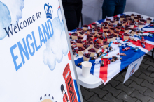 Biały baner z napisem ‘Welcome to England’ i grafiką chmury, korony, czerwonej budki telefonicznej i strażnika. Obok stół z flagą Wielkiej Brytanii, na którym leżą papierowe rożki ze słodyczami i pamiątki.