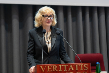 Kobieta, wiceminister zdrowia, z jasnymi włosami, w okularach i w ciemnym garniturze i koszuli w paski stoi za mównicą z napisem „VERITATIS SPLENDOR”. Przemawia, delikatnie uśmiechając się. Przed nią dwa mikrofony, obok butelka wody i szklanka. W tle szare zasłony i fragment czerwonego krzesła.