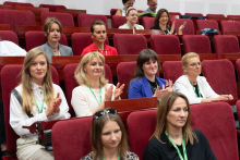 Grupa osób siedzi w czerwonych fotelach w audytorium lub sali konferencyjnej. Wszyscy mają identyfikatory na zielonych smyczach. Twarze zostały zamazane. W tle widoczne są kolejne osoby zwrócone w stronę sceny lub mównicy.