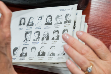 Zbliżenie na ręce trzymające kartkę z napisem „GRUPA IX” i 18 czarno-białymi zdjęciami portretowymi ułożonymi w trzech rzędach. Pod każdym zdjęciem widnieje imię i nazwisko. W tle widoczne są inne dokumenty i fragment stołu.