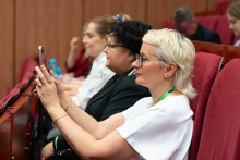 Kilka osób siedzi w rzędzie na czerwonych krzesłach podczass konferencji. Osoba na pierwszym planie z krótkimi blond włosami i w białej koszulce trzyma smartfon. W tle widoczne drewniane panele ścienne i inne osoby na widowni.