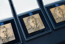 Trzy złote medale o kwadratowym kształcie, umieszczone w otwartych czarnych pudełkach z białym wnętrzem. Na każdym medalu widoczny jest relief z twarzą oraz wygrawerowany tekst po prawej stronie.