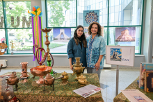 Wystawa poświęcona Iranowi. Na stole z zielonym obrusem w perski wzór ustawiono miedziane naczynia, dzbanki i miski. W tle plakaty z irańskimi zabytkami, tablica z napisem „IRAN” oraz książki i broszury. Za stołem stoją dwie młode kobiety, uśmiechają się do zdjęcia.