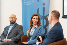 Three people in formal attire sit on brown chairs and hold microphones. In the background is a blue banner with the words “DEPARTMENT OF HEALTH SCIENCES” and a caduceus symbol.