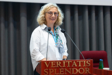 Kobieta, prodziekan, z jasnymi, kręconymi włosami, w okularach, stoi za mównicą z napisem „VERITATIS SPLENDOR”. Ubrana jest w białą koszulę i kolorowy szalik. Przed nią znajdują się dwa mikrofony. W tle widoczne są ciemne zasłony. Po prawej stronie, na stole obok mównicy, stoi butelka z wodą i kubek.