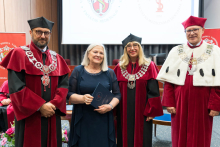 Cztery osoby w strojach akademickich, trzy w czerwonych togach z łańcuchami, jedna w białej todze. W centrum kobieta w granatowej sukience trzyma dokument. W tle logotypy uczelni i zasłony.