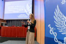 Kobieta stoi na scenie i trzyma mikrofon. W tle widoczny jest ekran z prezentacją zatytułowaną „Udar mózgu – problem kliniczny i społeczny w kontekście aktualnych rekomendacji – rola logopedy klinicznego”. Pod tytułem widnieją dane prelegentki: Izabela Domitrz, Klinika Neurologii WUM, Szpital Bielański w Warszawie. Po prawej stronie ekranu znajduje się logo z laską Eskulapa.