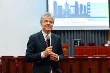 Mężczyzna - prorektor prof. Marek Kuch, w garniturze i krawacie stoi z mikrofonem w auli. W tle ekran z panoramą miasta. Widoczne są rzędy czerwonych krzeseł i mównica.