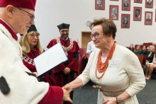 Kobieta w kremowej sukience i pomarańczowym naszyjniku odbiera dyplom od mężczyzny w czerwonym birecie i stroju ceremonialnym. W tle osoby w togach akademickich, publiczność oraz portrety na ścianie.