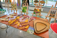 Stół z rękodziełami, takimi jak wachlarze, kapelusze i ozdobne maty. Na stole leży broszura z napisem „MALAYSIA”. W tle obrazy na sztalugach i dekoracje. Pomieszczenie jasne, z dużymi oknami.