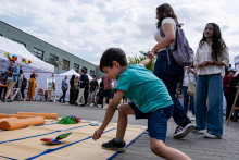 Chłopiec w zielonej koszulce i niebieskich spodenkach rzuca kolorowe woreczki na matę rozłożoną na bruku. W tle grupa ludzi i stoiska z białymi namiotami, na jednym z nich widoczne kolorowe balony.