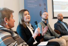 Four people sit on chairs during a panel discussion. A woman in a black jacket holds a microphone and gestures. Next to her a man in a gray suit, further away a man in a blue jacket. In the background is a blue banner that reads “UNIVERSITY OF MEDICINE DEPARTMENT OF HEALTH SCIENCES” and a screen with a presentation.