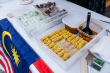 Stół z potrawami i flagą Malezji. Na stole trzy pojemniki: zielone kulki z białym proszkiem, kawałki ciasta czekoladowego oraz żółte naleśniki z miską czerwonego sosu i chochlą. Obok leżą drewniane pałeczki i plastikowe sztućce.