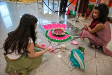 Dwie kobiety w tradycyjnych strojach indyjskich siedzą na podłodze i tworzą rangoli – dekorację z kolorowego proszku w kształcie kwiatu z różowymi płatkami, żółtym środkiem i białymi wzorami. Wokół leżą pojemniki z proszkiem i narzędzia.