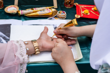 Dwie osoby uczestniczą w malowaniu dłoni henną. Osoba po lewej w tradycyjnym stroju z bransoletką trzyma dłoń, na której druga osoba aplikuje hennę złotą tubką. Na stole leżą stożki z henną, kolorowe pudełka i ozdoby.