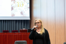 Kobieta z długimi blond włosami stoi w sali konferencyjnej, trzyma mikrofon. Ubrana w czarną bluzkę i ciemną marynarkę. W tle drewniana ściana i ekran z rozmytym obrazem oraz liczbą '10'. Na pierwszym planie fragment szarego fotela.
