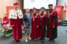 Pięć osób pozuje do zdjęcia grupowego. Cztery w czerwonych togach z łańcuchami, jedna w kolorowej sukience trzyma niebieską teczkę. W tle czerwone banery z herbem uczelni, ekran z logo i napisem „Universitas Medica”. Na podłodze bukiet kwiatów w białym koszu.