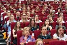 Aula z dużą grupą osób siedzących w czerwonych fotelach, zwróconych w stronę sceny poza kadrem. Uczestnicy mają identyfikatory na smyczach.