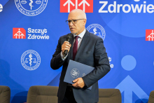 Mężczyzna w średnim wieku, rektor WUM, w ciemnym garniturze, białej koszuli i bordowym krawacie trzyma mikrofon oraz niebieską teczkę z logo Instytutu Ochrony Zdrowia. W tle niebieska ścianka z logotypami Instytutu i napisem „Szczyt Zdrowie”.