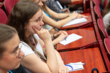 Grupa osób siedzi w rzędach na sali wykładowej. Część z nich notuje na papierze. Widać drewniane blaty stołów i czerwone krzesła. 