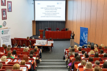 Sala wykładowa z uczestnikami siedzącymi na czerwonych krzesłach, skierowanymi w stronę podium i ekranu. Na ekranie definicja WHO dotycząca udaru mózgu. Po lewej stronie baner „Ogólnopolska Konferencja Logopedii Klinicznej”.