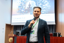 Mężczyzna - dr Dominik Ludwiczyński - w garniturze stoi z mikrofonem przed ekranem projekcyjnym. Na ekranie widoczny jest slajd z napisem „Człowiek 5.0” i „Przygotuj kompetencje na AI” oraz grafika medytującej sylwetki otoczonej ikonami technologii. Po lewej stronie mównica z napisem „VERITATIS SPLENDOR”.