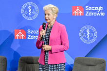 Kobieta w różowej marynarce i sukience w biało-granatowy wzór stoi z mikrofonem na tle niebieskiej ściany z logotypami „Instytut Ochrony Zdrowia” i „Szczyt Zdrowie”. Po obu stronach znajdują się dwa ciemne fotele.