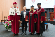 Cztery osoby stoją na tle drewnianej ściany i ekranu. Trzy w czerwono-czarnych togach z łańcuchami, jedna w garniturze trzyma czerwoną teczkę. Po lewej bukiet kwiatów, po prawej mównica z napisem „Veritas Splendor”.