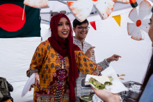 Młoda kobieta i młody mężczyzna stoją pod białym namiotem. Kobieta po lewej ma kolorową tunikę i bordowy szal na głowie, mężczyzna po prawej jasną koszulę z wzorem. Obie wskazują coś palcami. W tle flaga Bangladeszu i kolorowe girlandy. Na pierwszym planie talerz z jedzeniem.