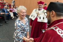  Starsza kobieta w sukience w niebiesko-białe fale i perłach ściska dłoń mężczyzny w czerwonym stroju akademickim. W tle drugi mężczyzna w czerwonej todze z futrzanym kołnierzem i łańcuchem rektorskim. Za nimi siedzą formalnie ubrani goście.
