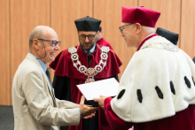 Starszy mężczyzna w jasnym garniturze odbiera dokument od osoby w białym płaszczu i czerwonym birecie. W tle osoba w czerwonym stroju akademickim z łańcuchem. Za nimi drewniana ściana panelowa.