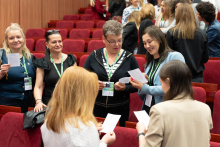 Grupa osób stoi w sali konferencyjnej z czerwonymi fotelami, prowadząc rozmowy i trzymając kartki. Większość ma identyfikatory na zielonych smyczach. W tle inne osoby rozmawiają lub przemieszczają się.