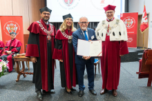 Cztery osoby w togach akademickich. Trzy w czerwono-czarnych togach, jedna w czerwonej todze z białym kołnierzem. Osoba w środku trzyma dyplom. W tle dwa czerwone banery z herbem uczelni i ekran z logo. Na pierwszym planie bukiet kwiatów na drewnianym stoliku.