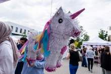 Pinata w kształcie jednorożca zawieszona na sznurku. Wykonana z białego papieru, z różowym rogiem i kopytami oraz grzywą i ogonem w kolorach niebieskim, żółtym i różowym. W tle uczestnicy wydarzenia plenerowego, namioty i budynki.