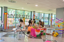 Kolorowo udekorowana przestrzeń wewnętrzna z dwoma stołami pełnymi dekoracji, w tym flagą Indii. W tle osoby przygotowujące ekspozycję. Na oknach napis „Diveristy x WUM” ozdobiony kwiatami i girlandami. Pomieszczenie otoczone barierkami, podłoga z płytek.